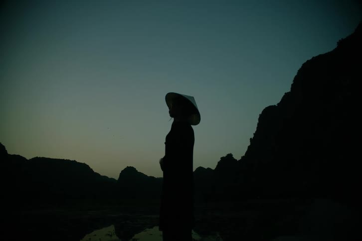 Person stands silhouetted near a reflective lake bordered by mountains in the evening