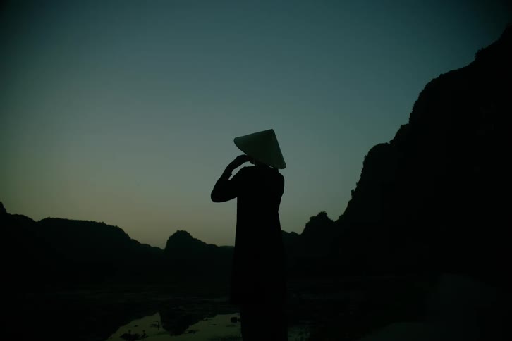 A person wearing a traditional conical hat stands still against a dark mountain landscape at twilight they eventually raise their hands to adjust the hat while a small flock of birds flies across the dim sky in the distance