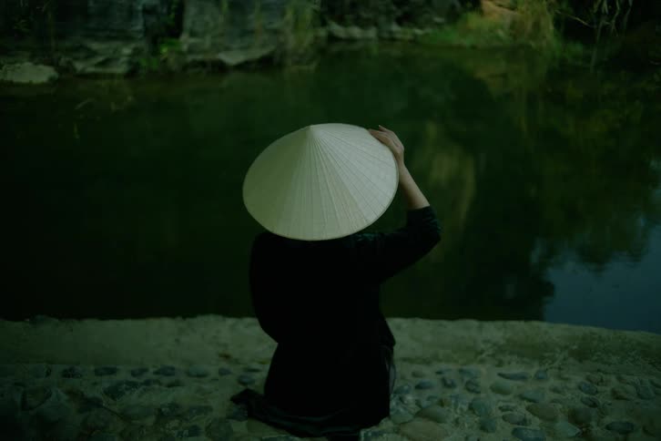 An asian woman holds onto her conical hat as she looks out upon a river