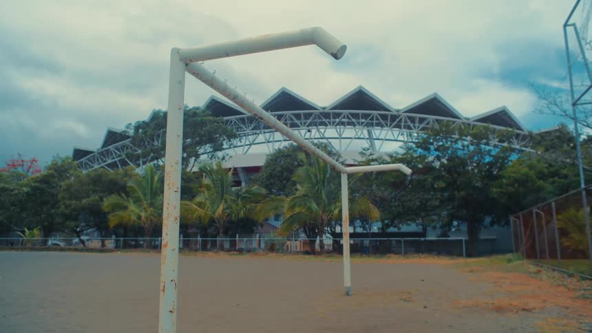 A view of a dirt field with two white metal goals in the foreground and a large stadium with a unique roof behind green trees under a cloudy sky