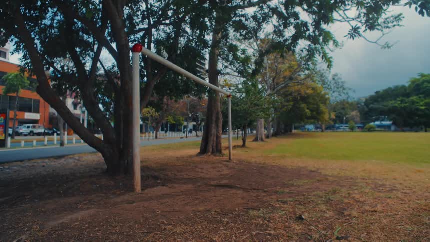A sturdy white goal post stands centered in a sundrenched urban park as leafy trees sway vigorously in the wind their branches dancing above a vast grass field that stretches toward a quiet city street lined with buildings