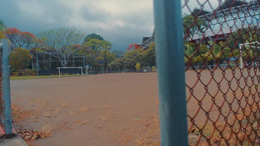 A view from behind a rusty chainlink fence shows a deserted dirt soccer field under a heavy grey sky with a large grandstand and lush trees in the background