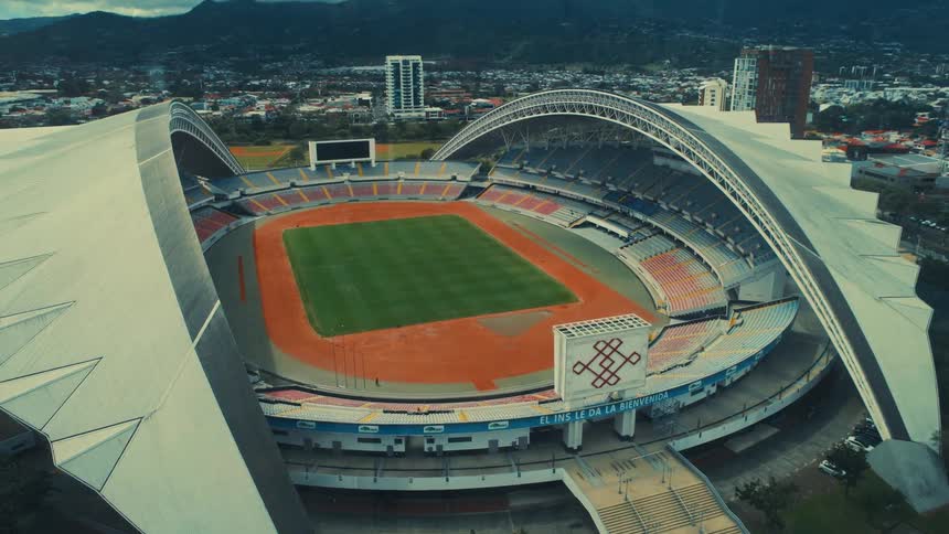 A wide aerial view shows a large modern sports stadium with a curved roof and empty colorful seats surrounding a green field and orange track with a city and mountains in the distance