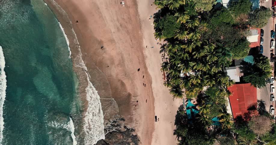 Aerial view of a neighborhood along the coast with cars on the streets and people walking on the beach as waves roll in to shore