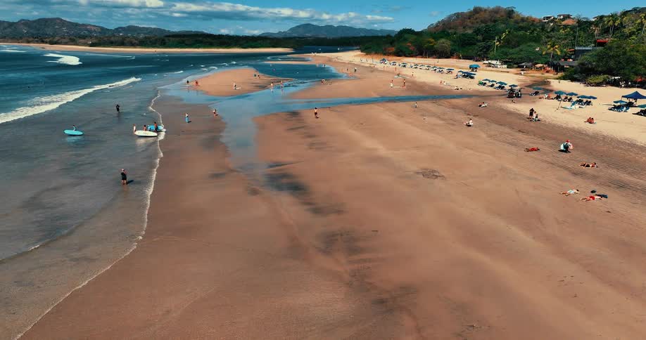 A wide tropical beach with waves crashing on the shore while people walk near a calm inlet and rows of blue umbrellas sit on the sand against a backdrop of lush green trees and distant mountains under a clear sky