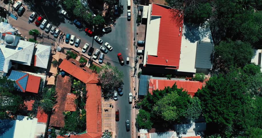 An aerial view shows cars driving along a paved road and through an intersection in a town with orange tiled roofs and green trees under bright sunlight