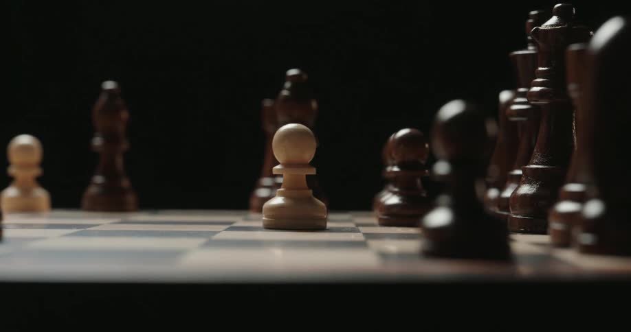 The video shows a closeup dramatic view of a hand moving a white pawn across a wooden chessboard the lowkey lighting emphasizes the texture of the pieces as the pawn is placed strategically among the dark opponents