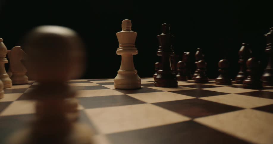 A close view shows a white king and a black king standing at the center of a wooden chess board surrounded by other pieces against a dark background