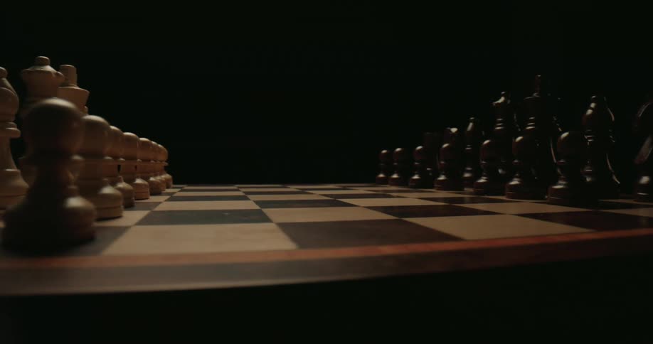 The camera performs a smooth lowangle tracking shot across a wooden chessboard moving from the lightcolored pieces toward the darkcolored onesthe scene is set against a pitchblack background with dramatic lighting that highlights the silhouettes