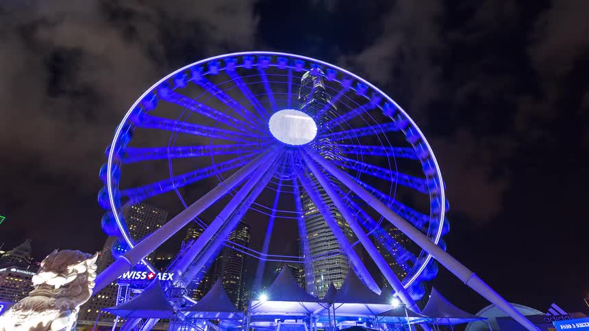 A large ferris wheel illuminated in blue rotates at night in a city with skyscrapers
