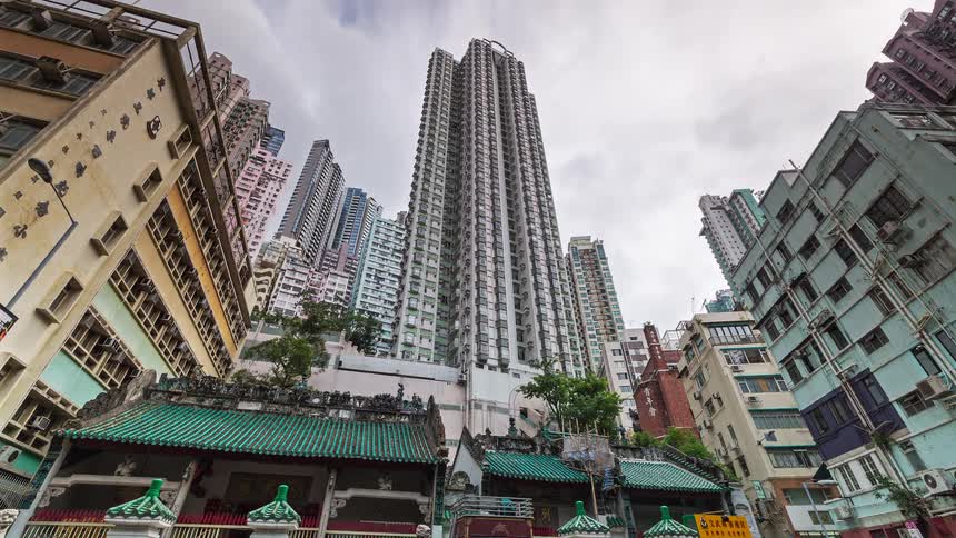 A view from below shows traditional buildings with green tiled roofs in the foreground against a backdrop of tall modern skyscrapers reaching into a grey overcast sky