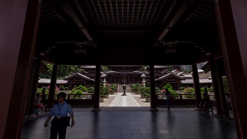 A view from a courtyard shows a large traditional wooden temple with curved roofs surrounded by manicured bonsai trees in stone pots under a blue sky with white clouds and modern skyscrapers in the background