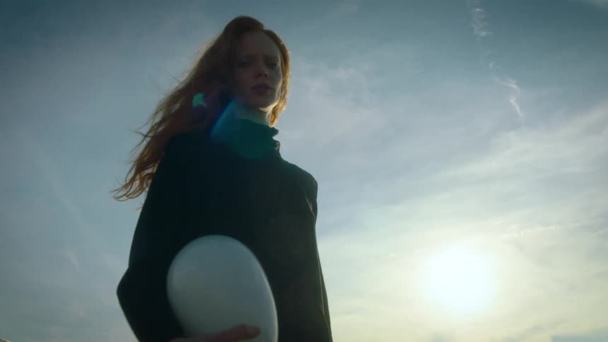 A view from below shows a woman with long red hair wearing black clothing as she looks around and runs against a blue sky with white clouds