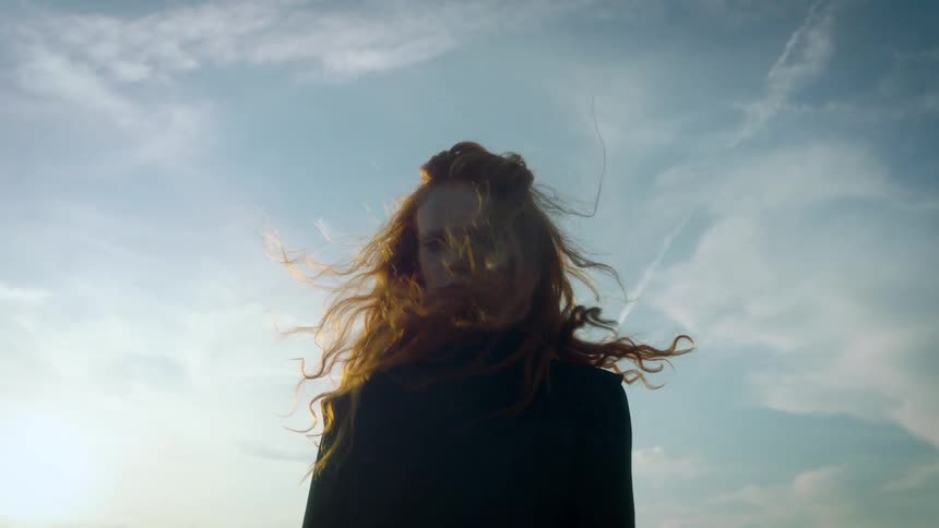 A woman with long red hair stands against a bright sky as the wind blows her strands across her face before she turns her back to the camera on a rooftop