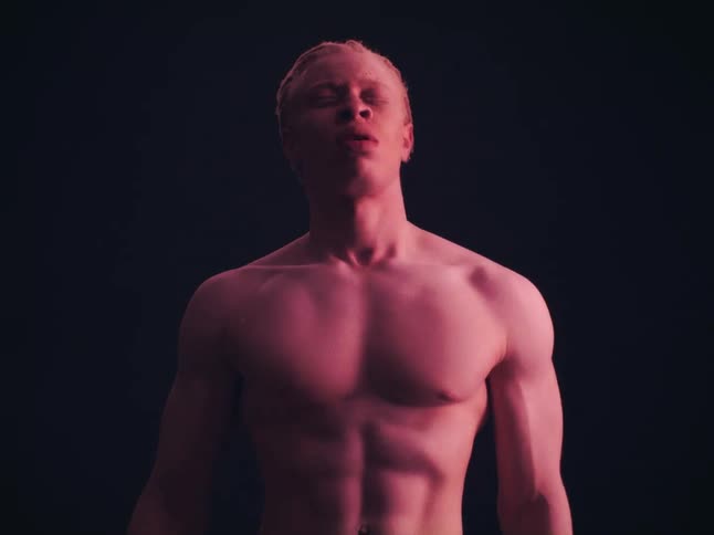 A shirtless man with pale skin and light hair stands against a dark background while red light glows from above as he keeps his eyes closed and his face tilted upward
