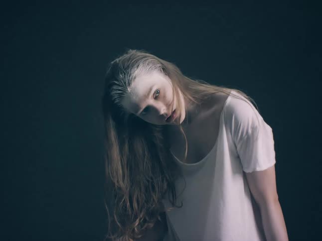 A young woman with pale white makeup and long messy hair leans toward the camera against a dark background while wearing a plain white tshirt in a moody studio setting