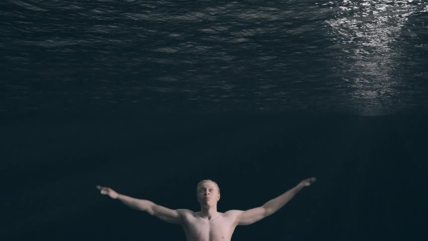 A view from below the surface of dark water shows a shirtless man with light skin and hair with his arms outstretched