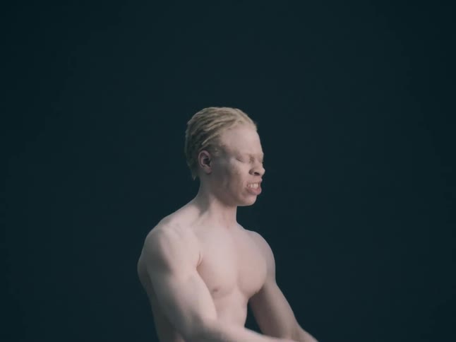 A close view shows a person with short blonde hair and white powder on their face as they move their head and arms quickly against a dark background