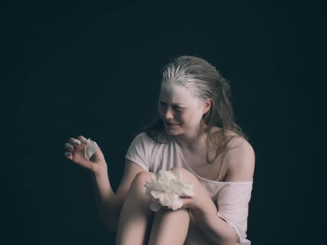 This video depicts a woman covered in white powder holding a flower with a distressed and emotional expression on her face as she sits in a dark setting she begins to tear the petals off the flower letting them fall to the ground