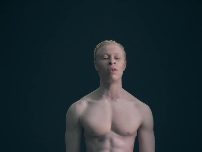 Albino black man standing up shirtless and talking while pacing and making gestures indoors