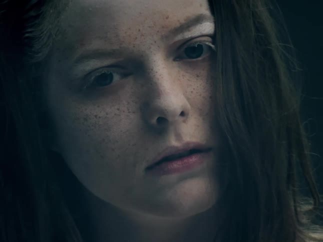 A close view of a woman with freckles and dark hair looks directly into the camera with a neutral expression in a dimly lit setting