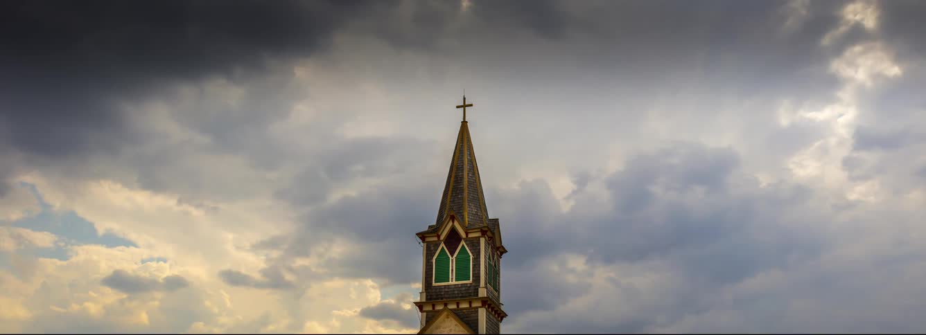 A weathered church spire with a golden cross stands prominently against a heavy overcast skydark clouds move swiftly behind the steeple creating a dramatic and moody atmosphere as the light shifts