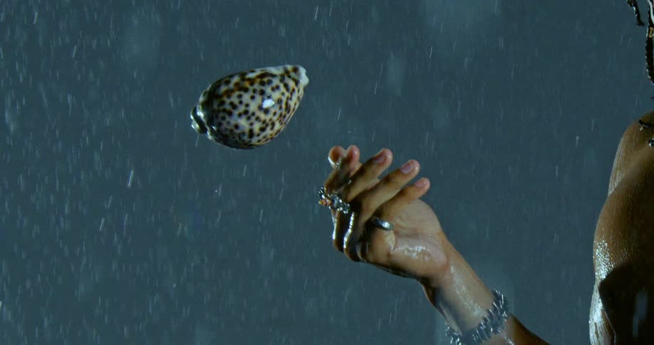 A view from the side shows a person wearing rings and a chain bracelet tossing a spotted seashell into the air while rain falls against a dark background