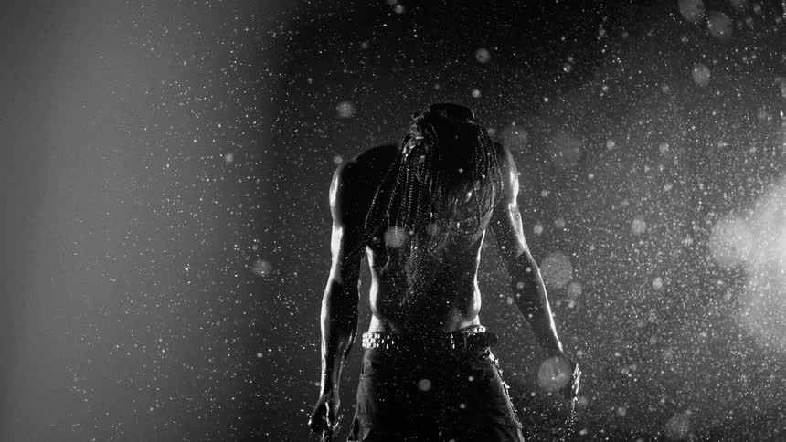 A shirtless man stands in dramatic blackandwhite lighting as water droplets fall around him creating a powerful atmospheric scene
