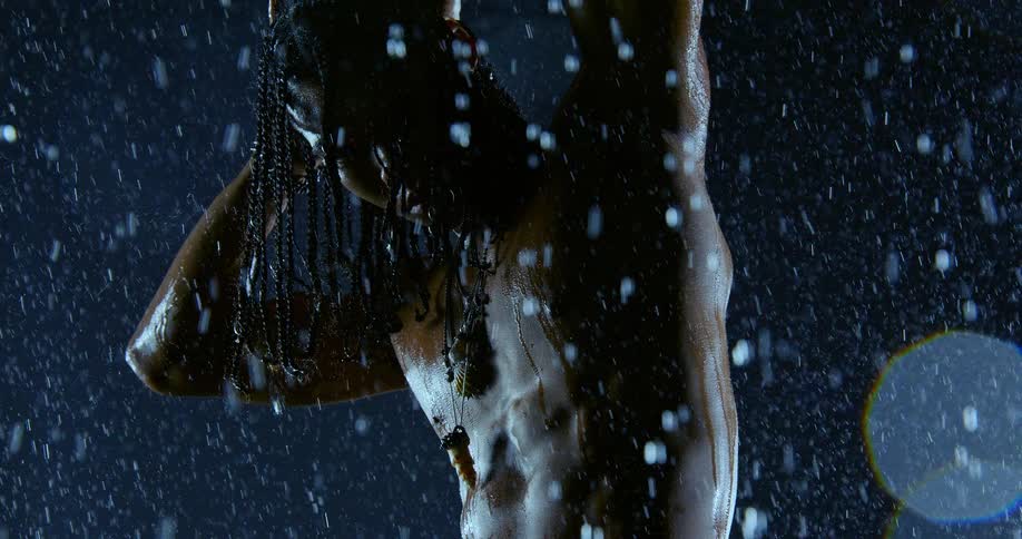 A person with long braided hair and jewelry on their face is seen in a blue light while water droplets fall and glisten against a dark background