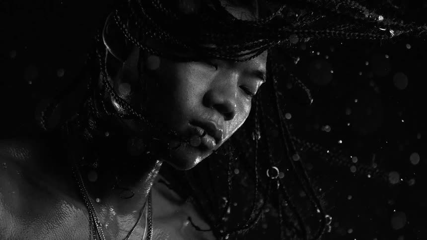 A man with long braids shakes his head in slow motion as water droplets fly through the air against a dark background in a high contrast black and white studio shot