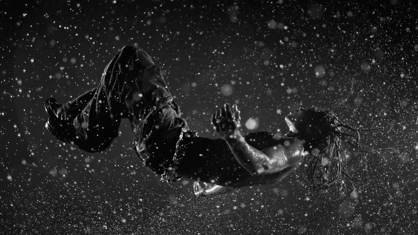 A shirtless man with braided hair appears suspended midair in dramatic blackandwhite lighting as water droplets swirl around him