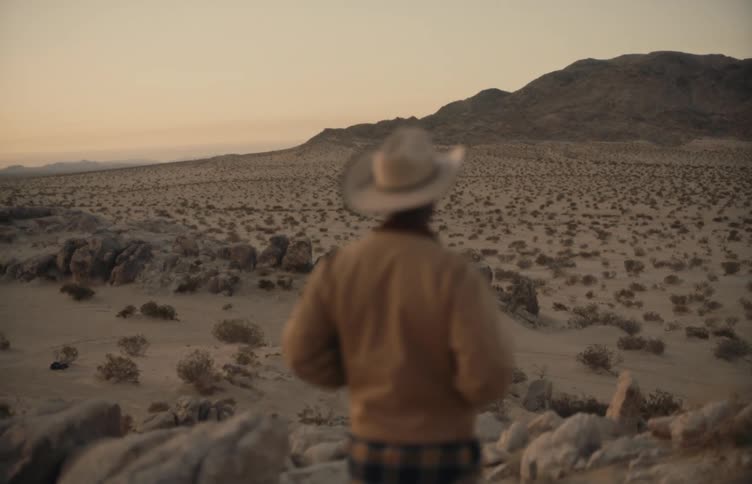Man wearing a hat observes a desert landscape with mountains at dusk