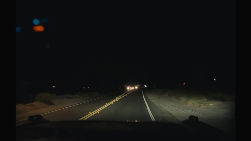 A dark rural road stretches ahead at night illuminated by headlights as distant cars approach in the opposite lane