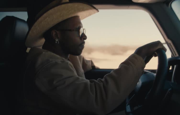 A man wearing a cowboy hat and sunglasses drives a vehicle through a sundrenched landscapethe camera remains steady in a sideprofile shot as he keeps his eyes on the road and hands on the wheel