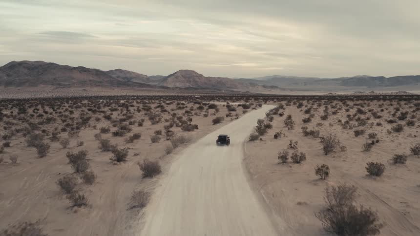 Vehicle drives towards the mountains on a dirt road passing through the desert on a cloudy day