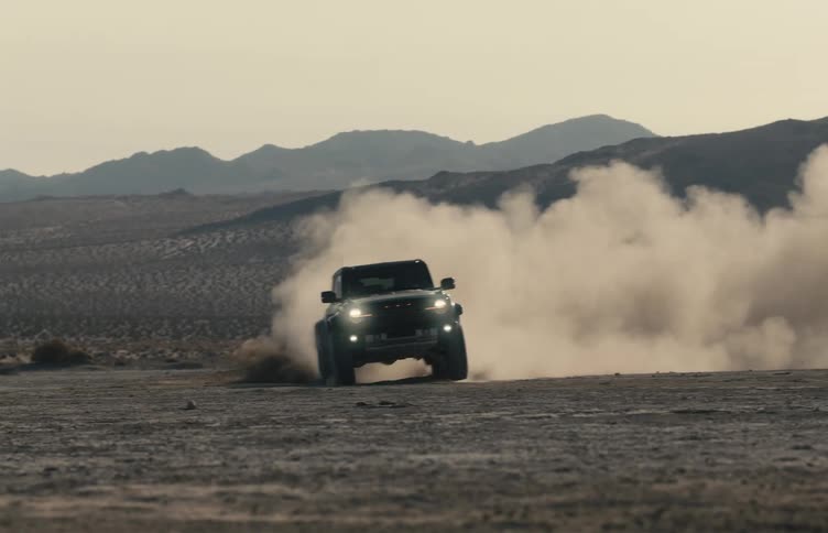 Utility vehicle moves across the desert raising a cloud of dust with mountains in the background