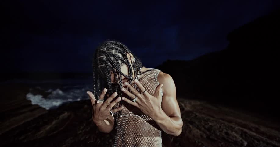 A person with braided hair and silver rings performs expressive hand movements while wearing a shimmering mesh top the setting is a dark dramatic coastline at night with crashing waves visible in the background