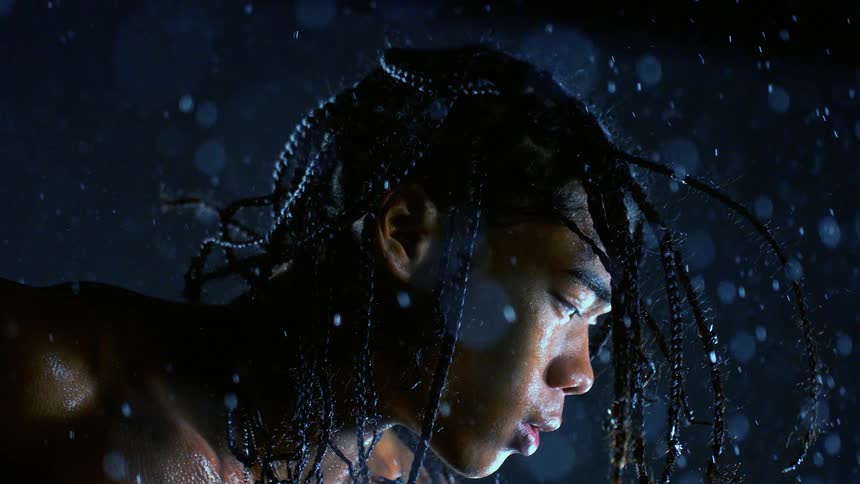 Man with braids under heavy rain with face sidelit against a dark background