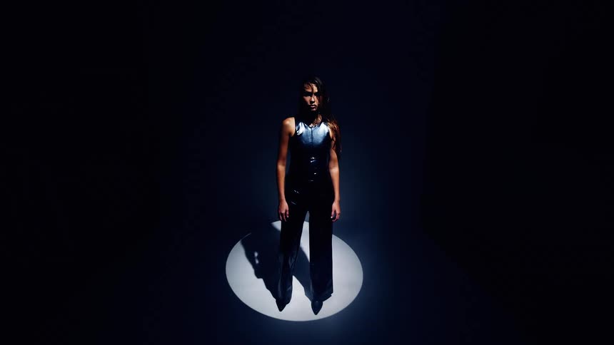 A young female model wearing a black jacket with braided hair stands in a spotlight on a stage striking a pose