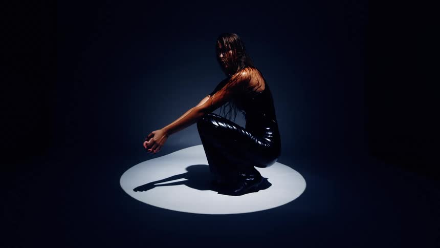 A woman with wet hair and dark glossy clothing crouches within a bright circular spotlight the surrounding environment remains in deep shadow focusing all attention on her silhouette and pose
