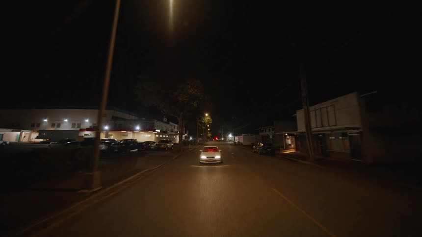 A silver sedan drives down a dark industrial street at night while passing parked cars and low buildings under the glow of overhead street lights