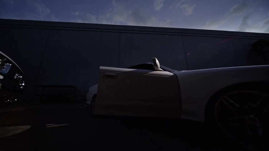 A person gets out of a white car and looks back while standing in a parking lot at night with other vehicles and a tall building in the background