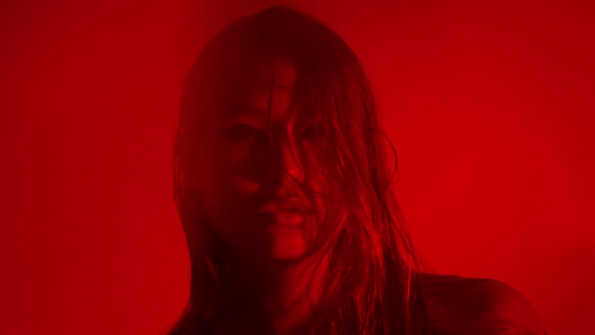 A woman with wet hair stares intensely toward the camera while bathed in a deep saturated red light her face fades in and out of a heavy blur creating a moody and atmospheric visual effect