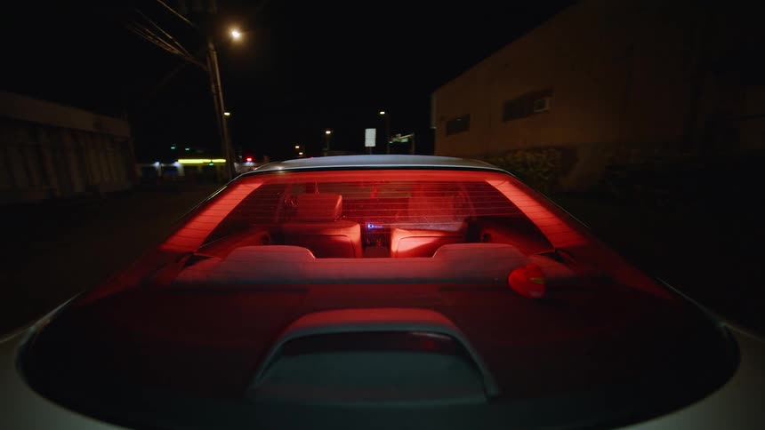 A view from behind a white car shows a nicty at night with utility poles and buildings