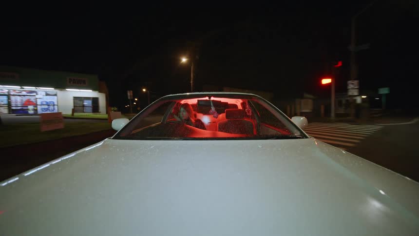 Inside the vehicle two people are visible through the windshield which is illuminated by a vibrant red glow from the dashboard and interior lights as the car moves it passes a pawn shop and several streetlights that create a sense of motion against th