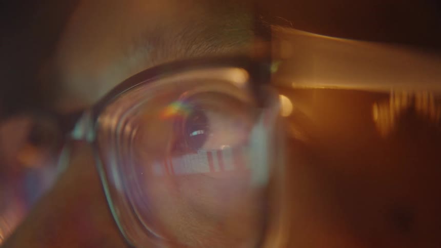 An extreme closeup of a persons eye behind glasses reflects warm light and colorful glare across the lens