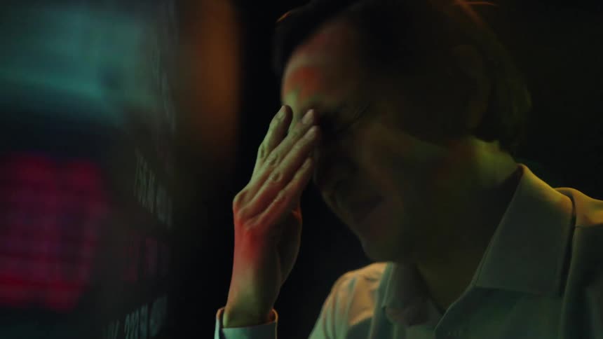 A man wearing glasses and a light colored shirt rubs his face and forehead in frustration while looking at a large screen displaying scrolling red and green stock market data in a dark room