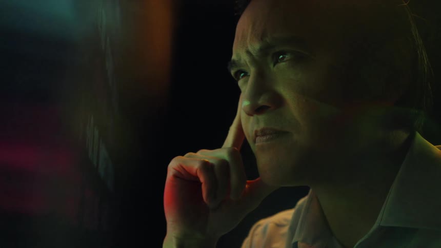 A man in a white shirt observes a digital stock ticker board and touches his temple