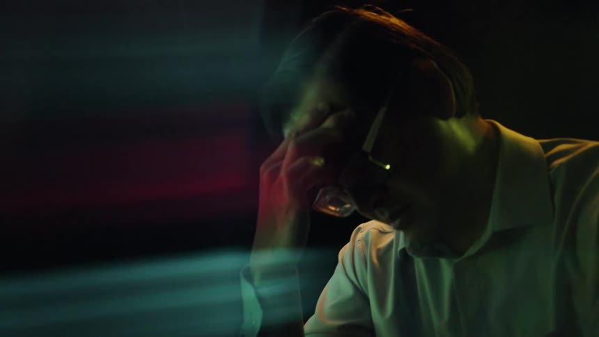 A man wearing glasses sits in dramatic colorful lighting from a screen with his hand pressed to his forehead appearing deep in thought or stressed