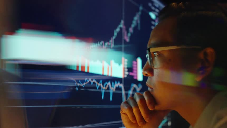 Man wearing glasses observes financial charts on a screen with his hand on his chin